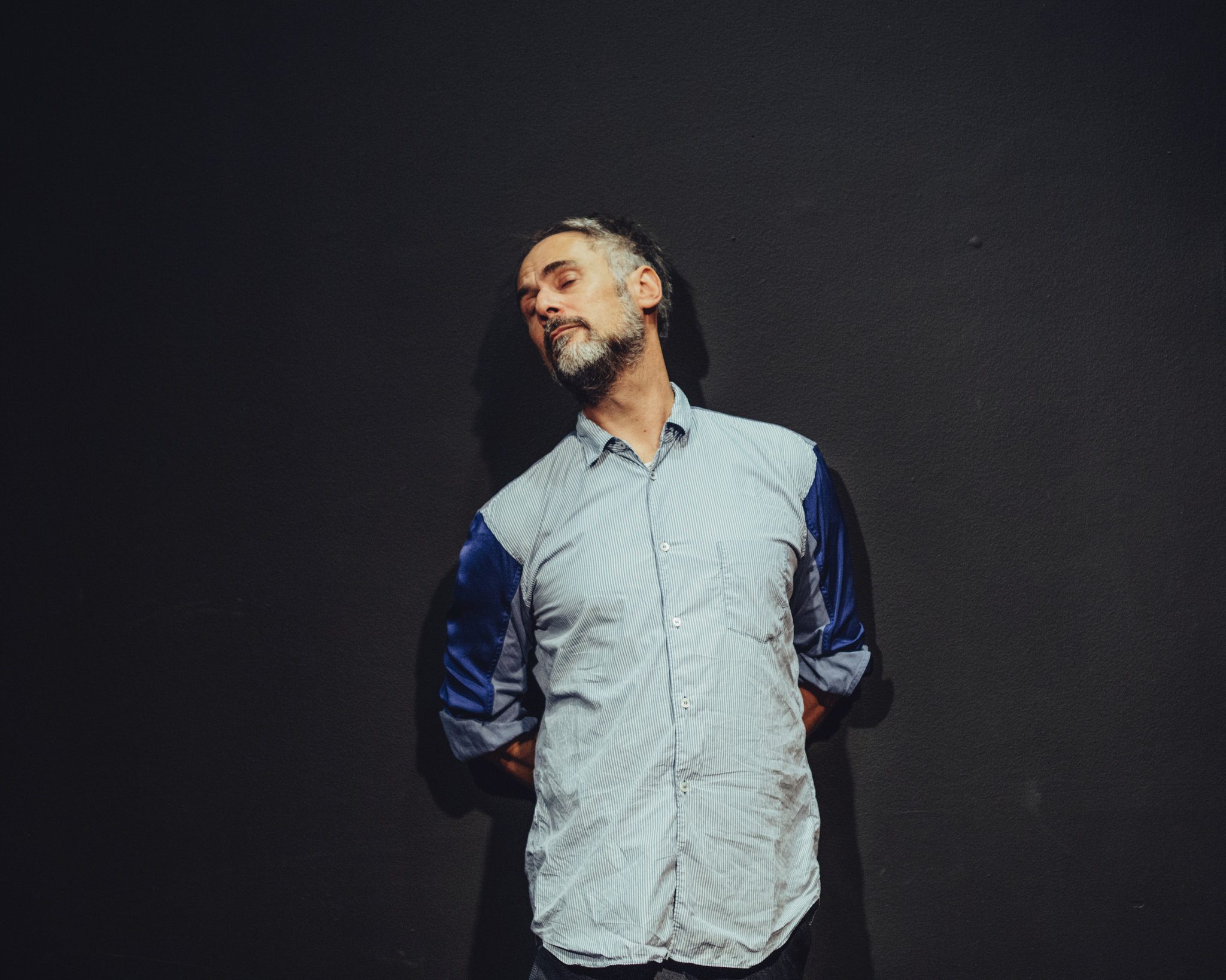 A man with a beard wearing a light blue shirt stands against a dark wall, tilting his head to one side with his hands behind his back.