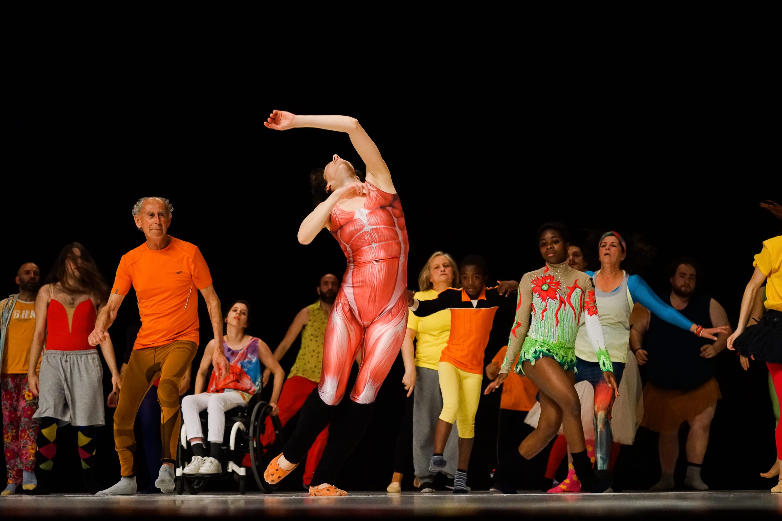 A diverse group of people, including a wheelchair user, dance energetically on stage. They wear colorful, eclectic outfits, expressing joy and movement against a dark background.