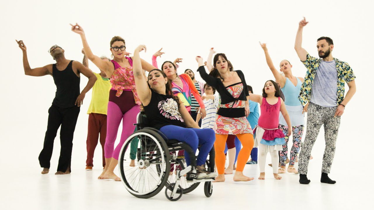A diverse group of people, including a woman using a wheelchair, dance together in colorful clothes against a plain white background.
