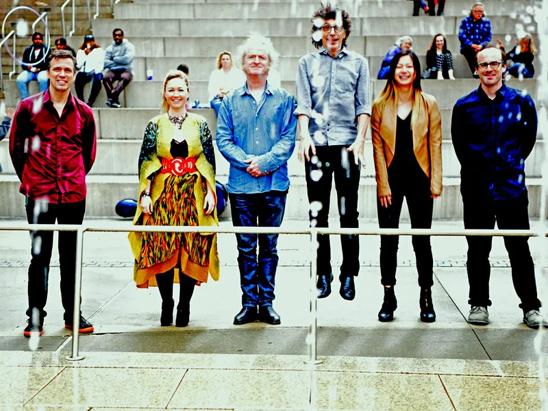 Five people stand side by side in front of stadium steps, some smiling; one person appears to be jumping. There are water droplets in the foreground and several people seated on the steps behind them.
