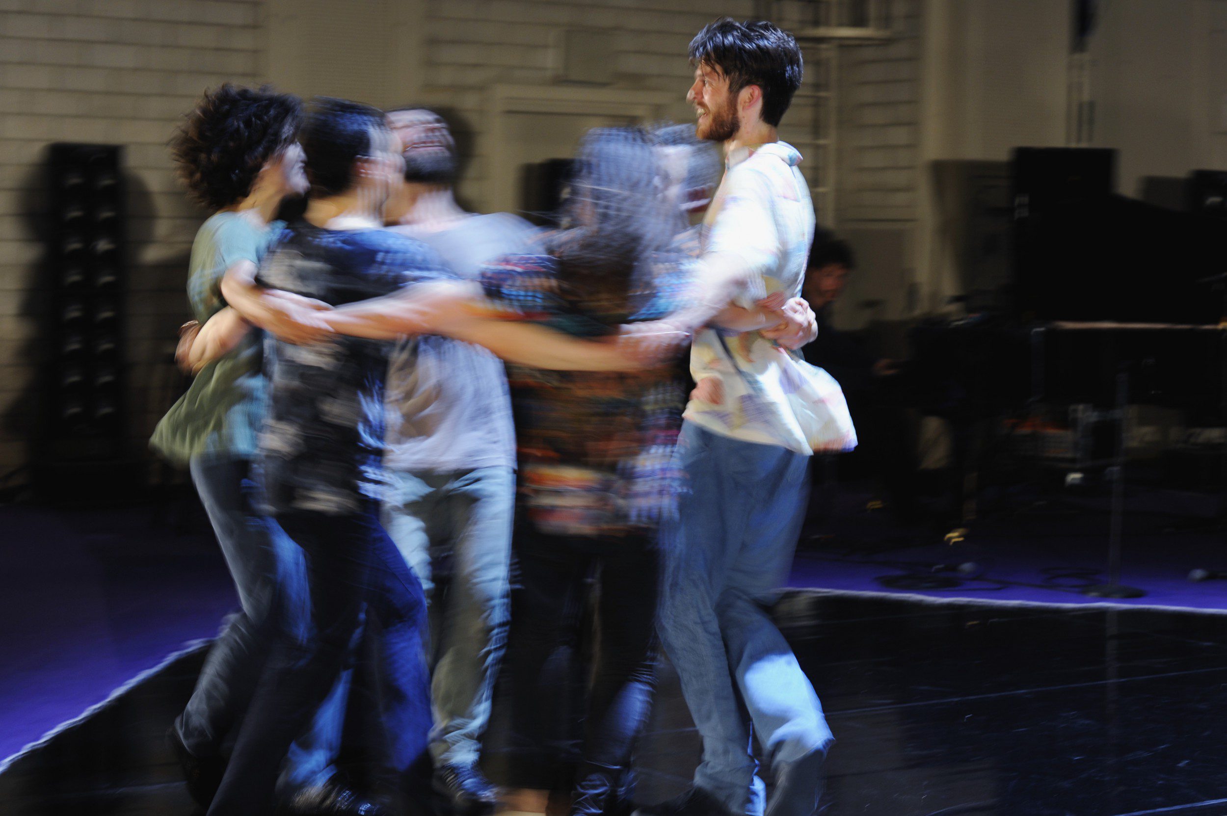 Six people are holding each other in a circle and spinning, causing a motion blur effect. They appear to be dancing or enjoying an energetic moment together indoors.