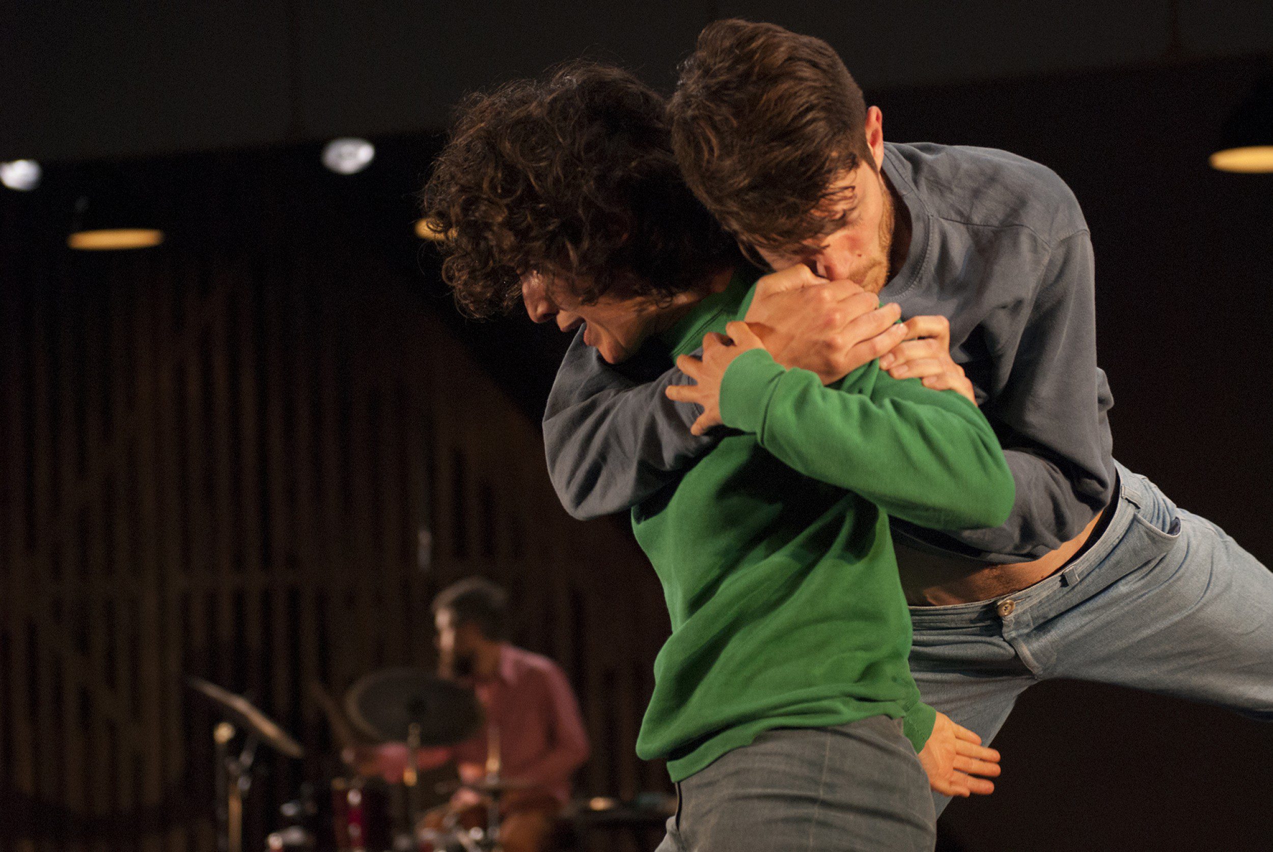 Two people embrace closely in the foreground, appearing to dance or perform. One person in a green shirt lifts the other, who wears blue. A drummer plays in the dimly lit background.