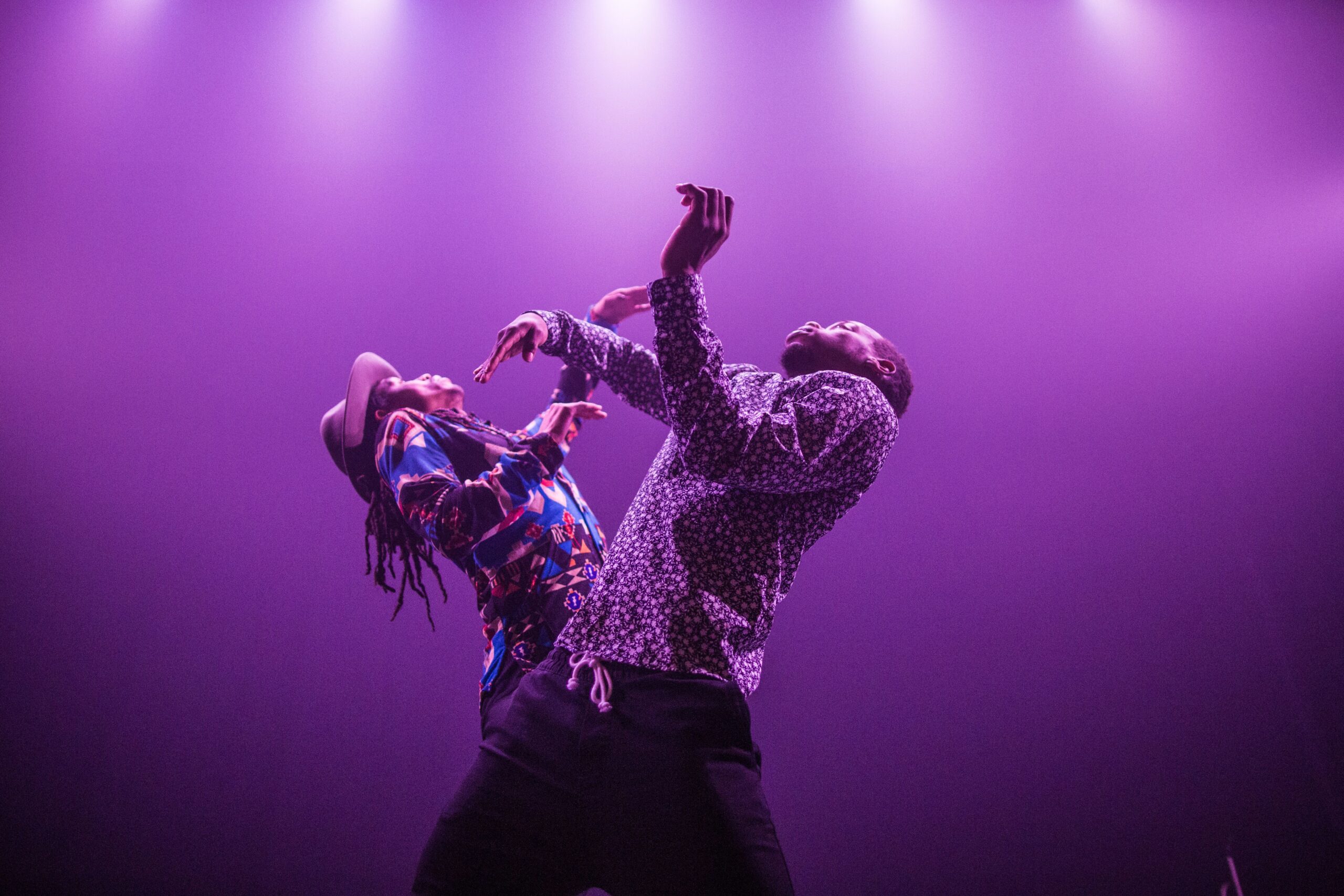 Two people dance dramatically under purple stage lights, their bodies arched and arms raised expressively. The colorful lighting and dynamic poses create an energetic, artistic atmosphere.