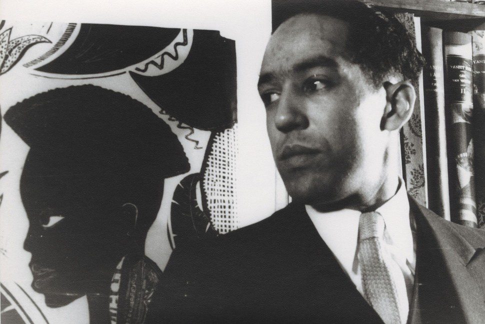 A man in a suit stands beside a painted profile of a woman with an Afro hairstyle; shelves with books are visible in the background. The image is in black and white.