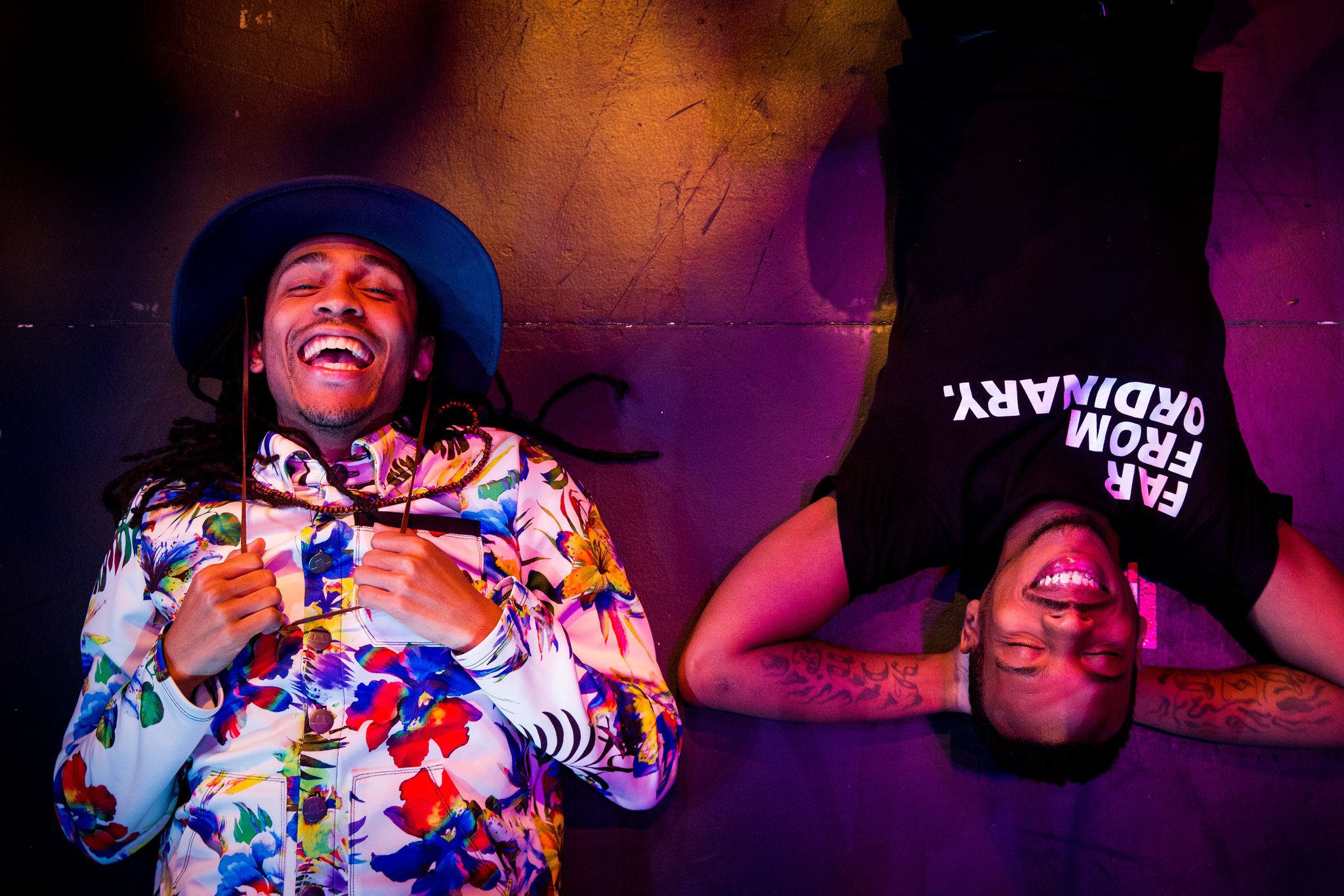 Two people lie on the ground, smiling and laughing. One wears a floral jacket and blue hat; the other wears a black shirt that reads “FAR FROM ORDINARY.” Colorful lighting creates a joyful atmosphere.