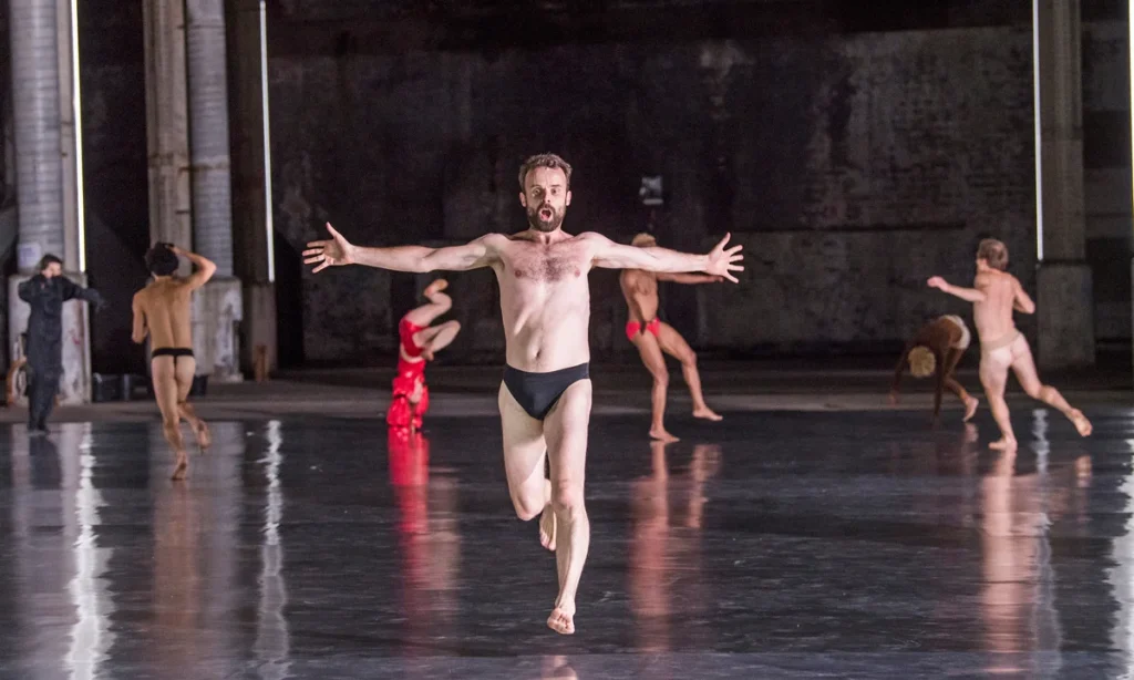 A man in black briefs runs toward the camera on a shiny stage, arms outstretched, while several other dancers move in various poses in the background.