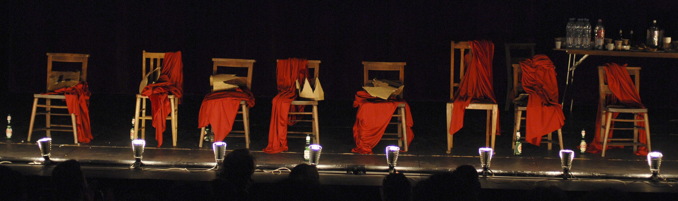 Seven wooden chairs draped with red fabric are onstage, each with a bottle at its base. A crown and papers sit on some chairs. A table with bottles and glasses stands at the right. Stage lights illuminate the scene.