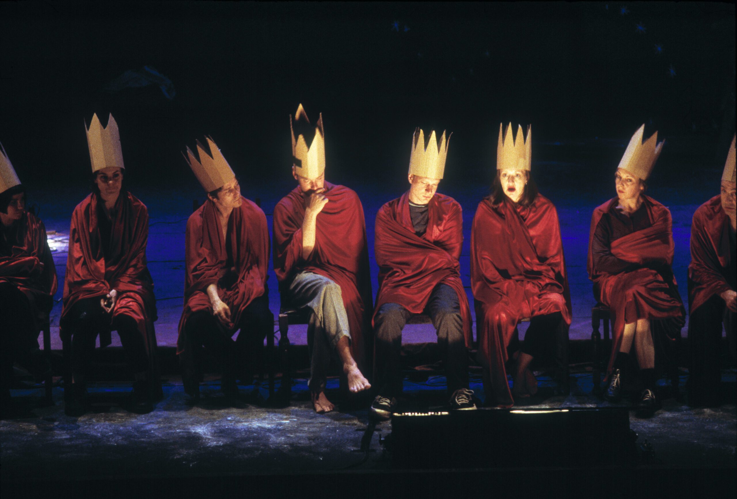 Eight people sit in a row on stage, each wearing a tall paper crown and wrapped in red fabric, bathed in blue and purple lighting, facing forward with various expressions.