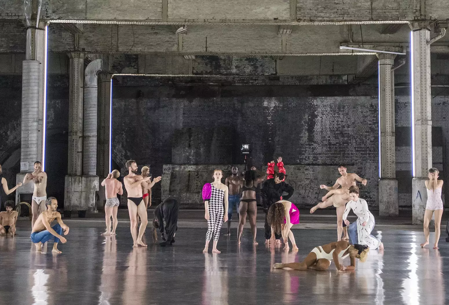 A group of dancers in varied poses and costumes perform on a shiny floor in an industrial space with brick walls, pillars, and minimal lighting.