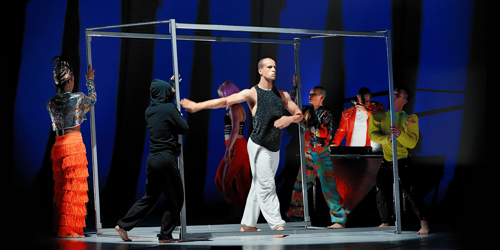 Six dancers perform onstage with a large metal frame, wearing colorful and varied costumes against a blue-lit background. The scene conveys dynamic movement and collaboration in contemporary dance.
