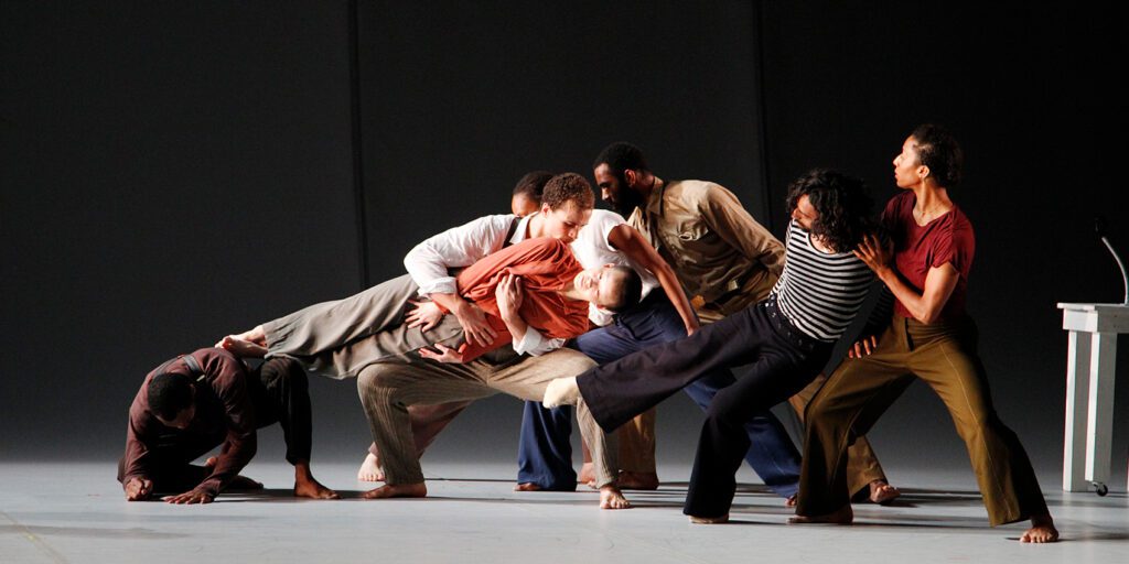 Seven dancers in varied poses support a person leaning horizontally across them. The group is on a bare stage, wearing loose clothing in earthy tones, with a desk visible at the right edge.