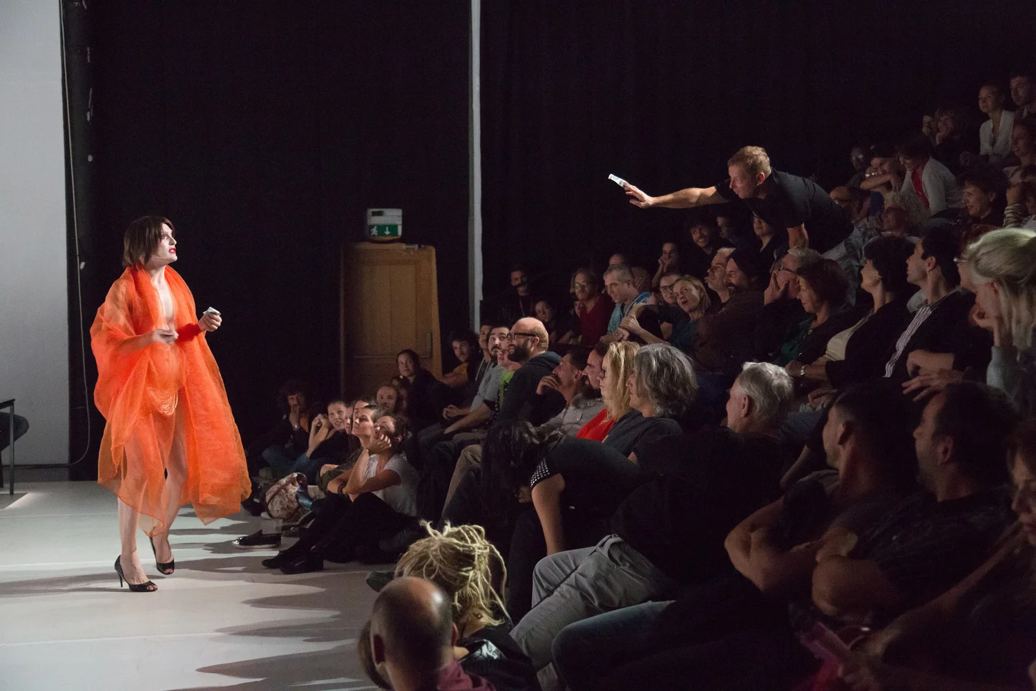A performer in an orange outfit stands on stage before a seated audience. A man in the crowd leans forward, extending a phone or device toward the performer. The audience looks engaged and attentive.