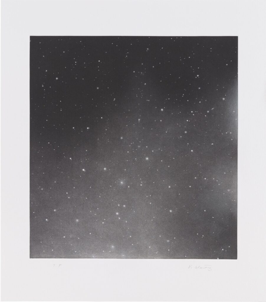 A black and white image showing a dense field of small, bright stars scattered across a dark sky. Some stars are more prominent, creating a sense of depth and vastness in the night sky.