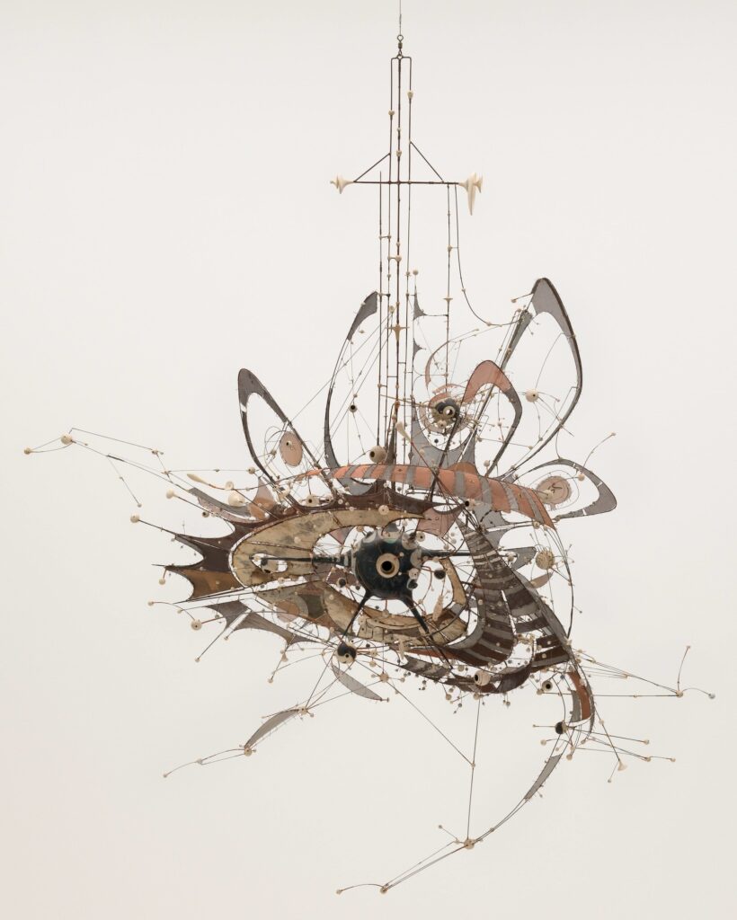A hanging kinetic sculpture made of metal rods, wires, and gears, forming an abstract, intricate, and airy structure with loops and protruding elements against a plain white background.