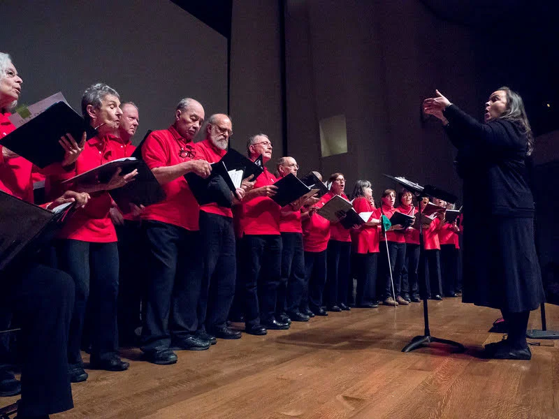 A choir, mostly older adults in red shirts, stands on stage singing while a woman conducts them. The scene is indoors with stage lighting.
