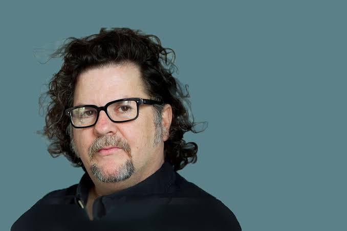 A man with curly dark hair, glasses, and a goatee, wearing a black shirt, poses against a plain teal background.