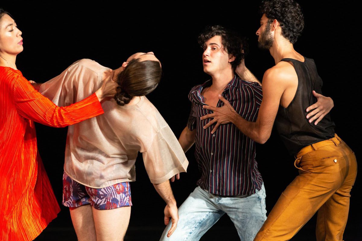 Four people perform an expressive dance. One person in red supports another, who leans back dramatically, while two others, in striped and black shirts, interact closely. The background is dark, emphasizing their poses.