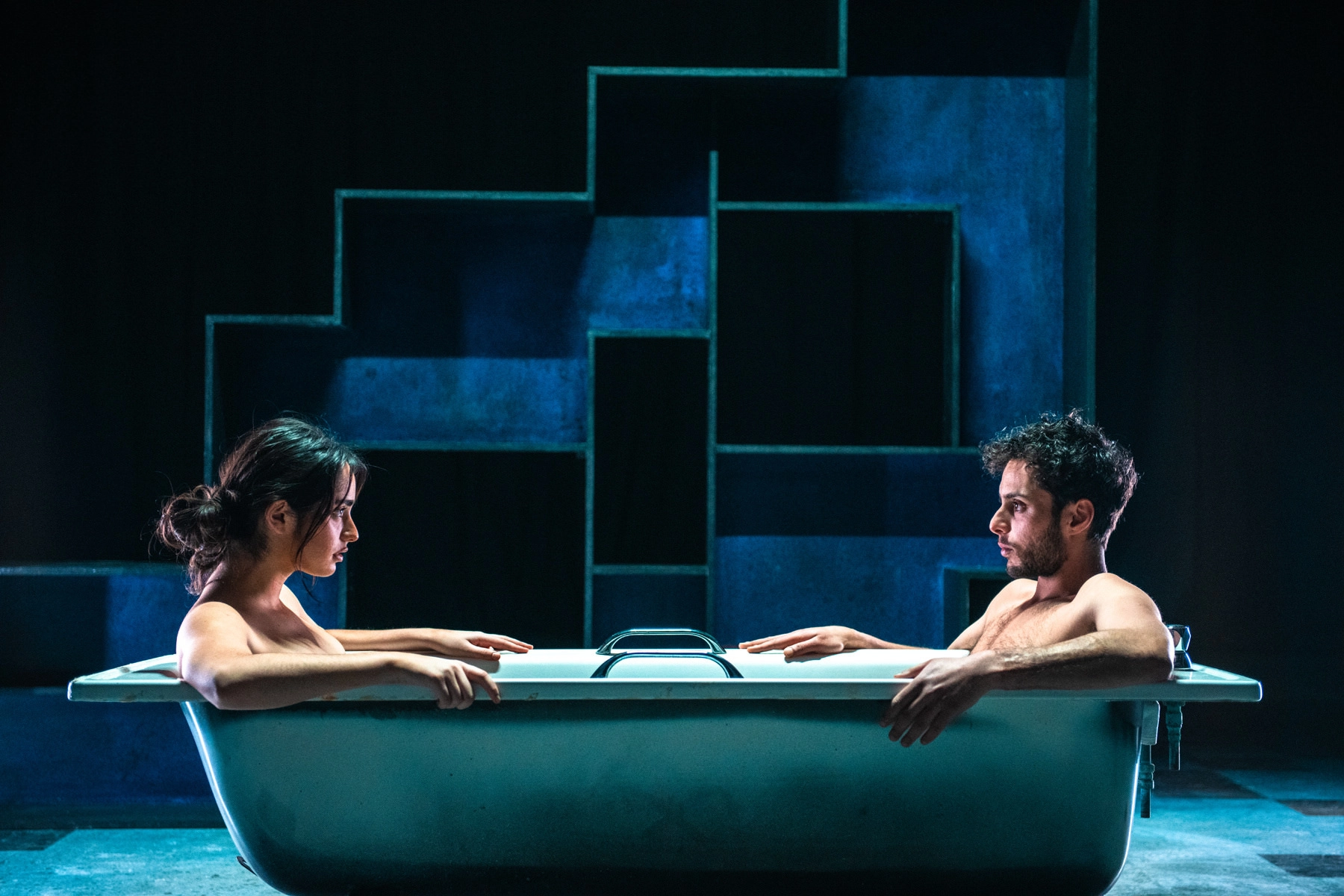Two people sit facing each other in a bathtub on stage, lit with dramatic blue lighting. Geometric set pieces form a backdrop, suggesting a theatrical or dramatic performance.