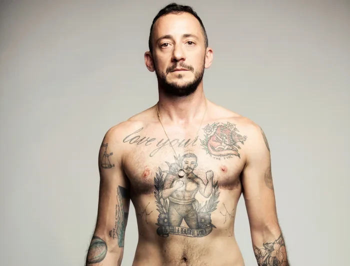 A man with tattoos, wearing red shorts and yellow boxing gloves, stands facing the camera against a plain background.