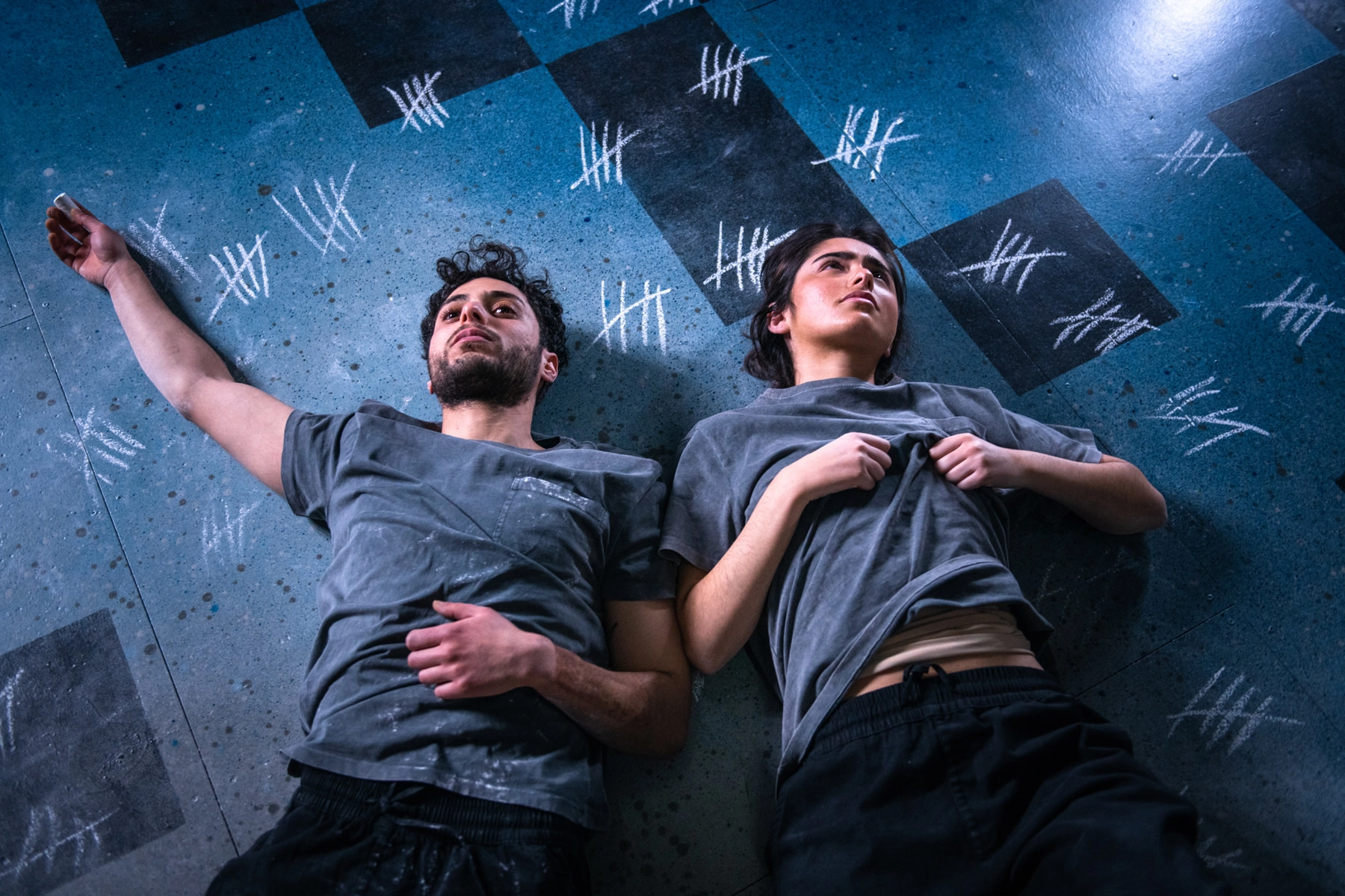 Two people lie on a blue floor covered in chalk tally marks, both wearing gray shirts and dark pants, looking up with tired expressions.