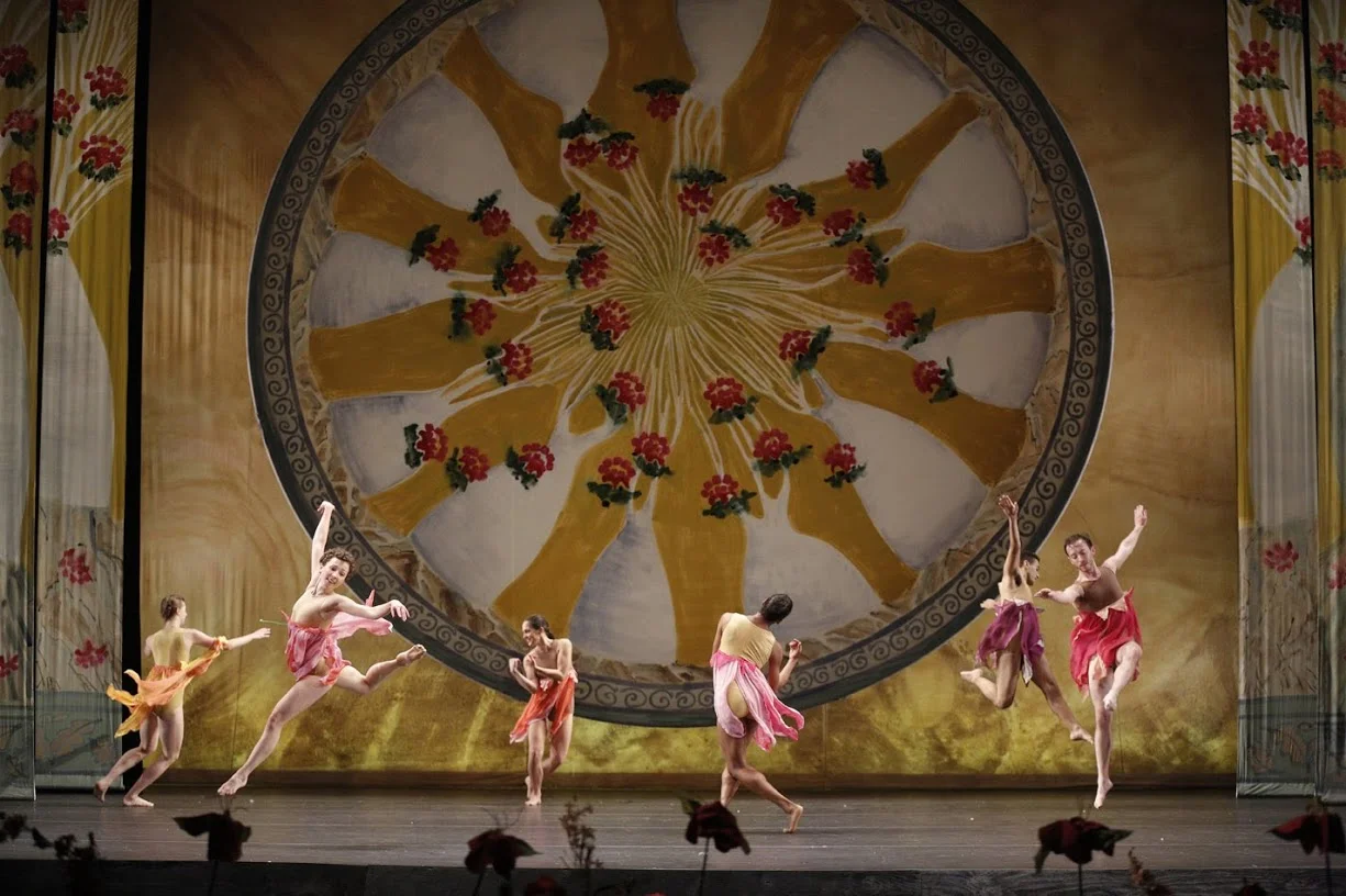 Dancers in colorful costumes perform on stage in front of a large, ornate backdrop featuring a circular floral and ribbon design.
