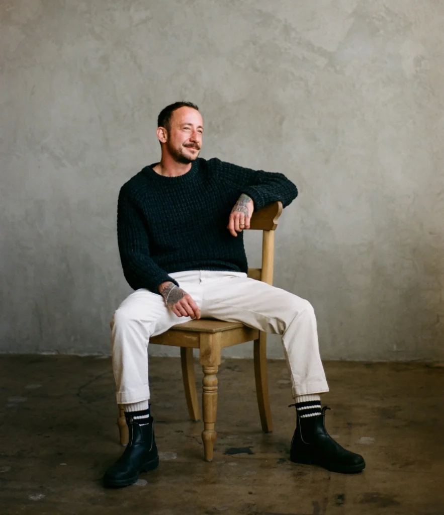A man with short hair and a beard sits on a wooden chair, wearing a dark sweater, light pants, and black boots, against a plain, textured wall.