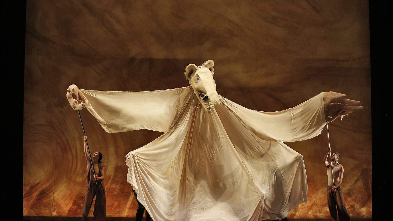 Three performers manipulate a large, flowing white puppet with a bear-like head and extended arms on a dramatic, earth-toned stage.