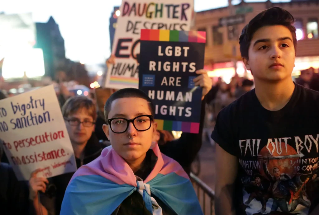 A group of people at a protest hold signs supporting LGBT rights, including one that reads “LGBT Rights Are Human Rights.” One person is draped in a trans pride flag.