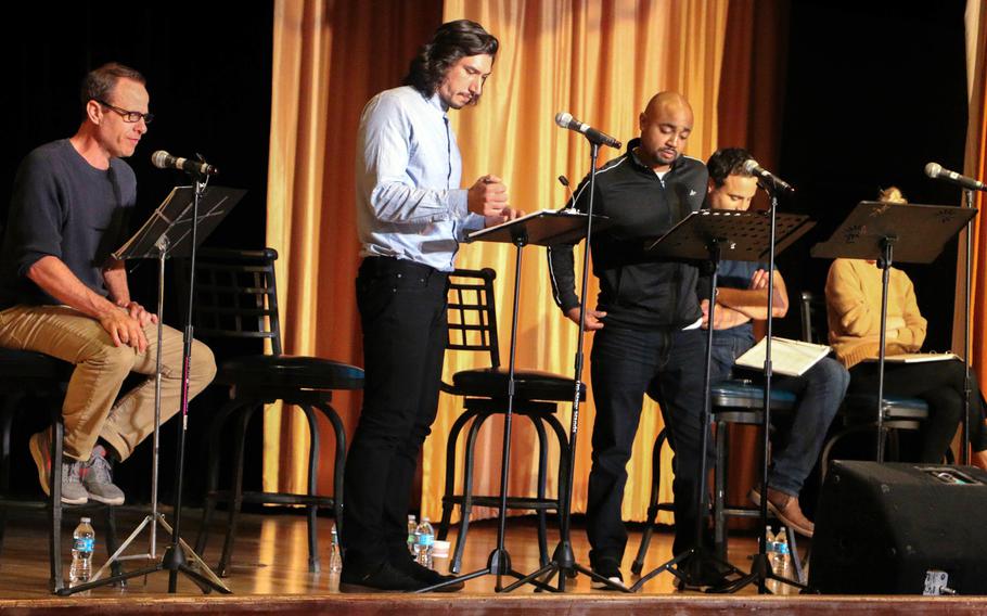 Five people participate in a staged reading on a theater stage, standing or sitting at music stands with scripts and microphones. Bottles of water are on the floor, and gold curtains hang in the background.