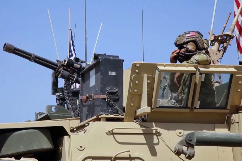 A soldier in camouflage gear sits in an armored vehicle, wearing a helmet with an American flag patch and sunglasses. A mounted machine gun and various antennas are visible, with a clear blue sky in the background.