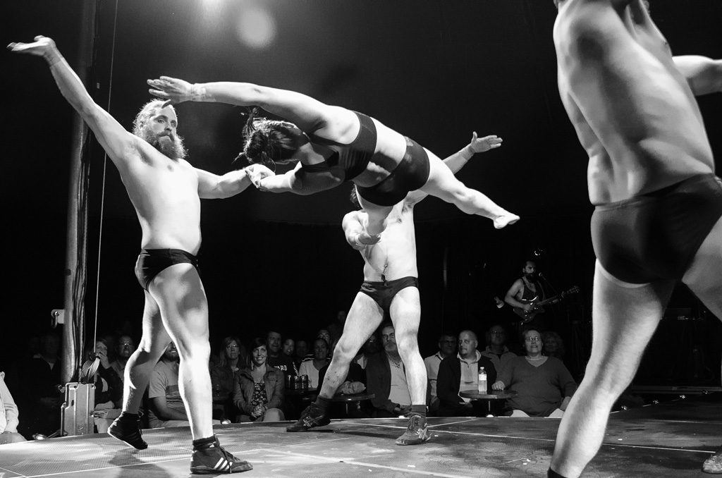 Four acrobats in dark briefs perform on stage; one acrobat is airborne, supported by another’s hand, as two more pose nearby. The audience watches the energetic act under dramatic stage lighting.