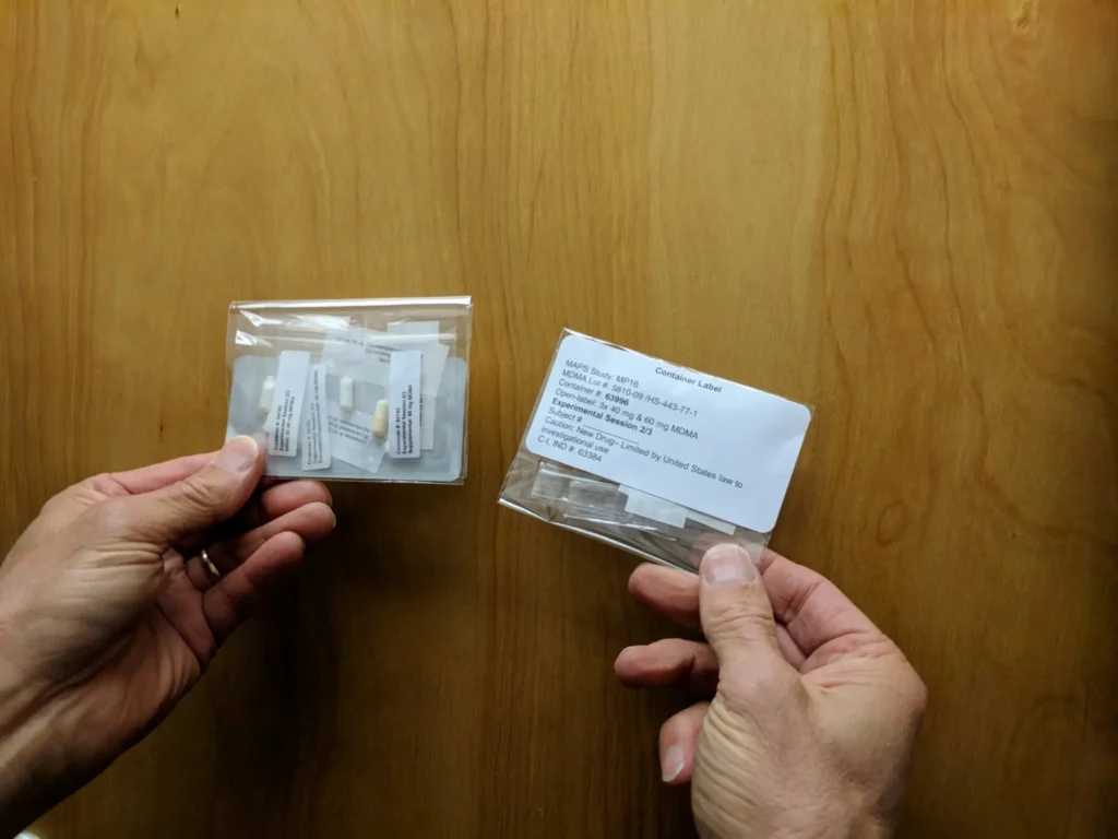Two hands holding small plastic bags containing labeled medical or laboratory vials or tubes, one bag is turned to show a white contents label with printed text. The background is a wooden surface.