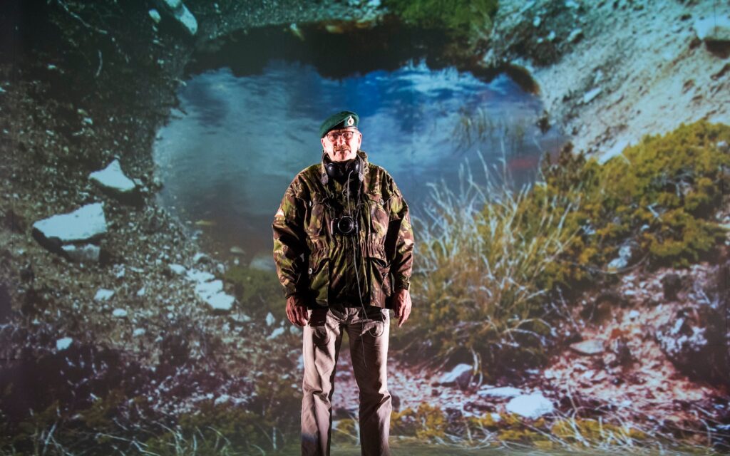 A man in a camouflage jacket and green beret stands in front of a large projected image of a pond and rugged outdoor landscape.