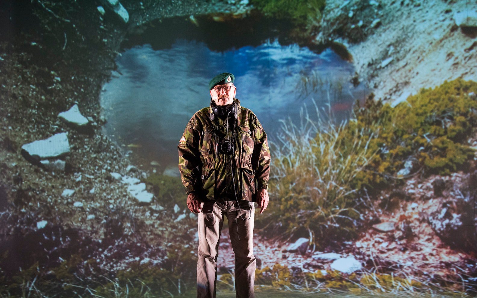 A man in a camouflage jacket and green beret stands in front of a large projected image of a pond and rugged outdoor landscape.