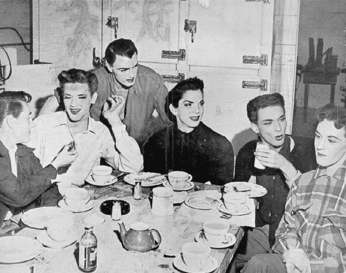 Six people with styled hair and makeup sit around a table set with teacups and a teapot, engaging in conversation and smiling, in a black-and-white vintage setting.