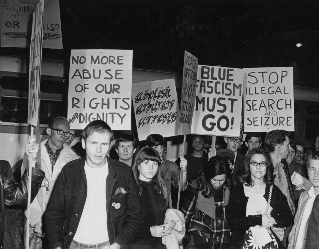 A black-and-white photo of protesters holding signs with messages such as “No more abuse of our rights and dignity” and “Blue fascism must go!” during a night demonstration.
