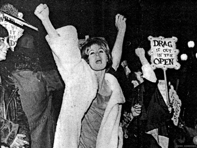 Black and white photo of people protesting at night, with one person in front raising their arm. Others hold signs, including one that reads “DRAG IT OUT IN THE OPEN.” The scene is lively and expressive.