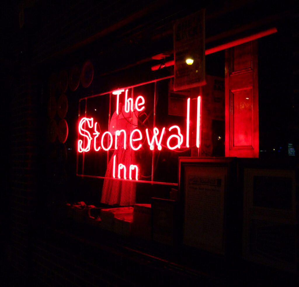 A neon sign glows red in a dark window, reading “The Stonewall Inn.” Some framed pictures and a dress are faintly visible inside.
