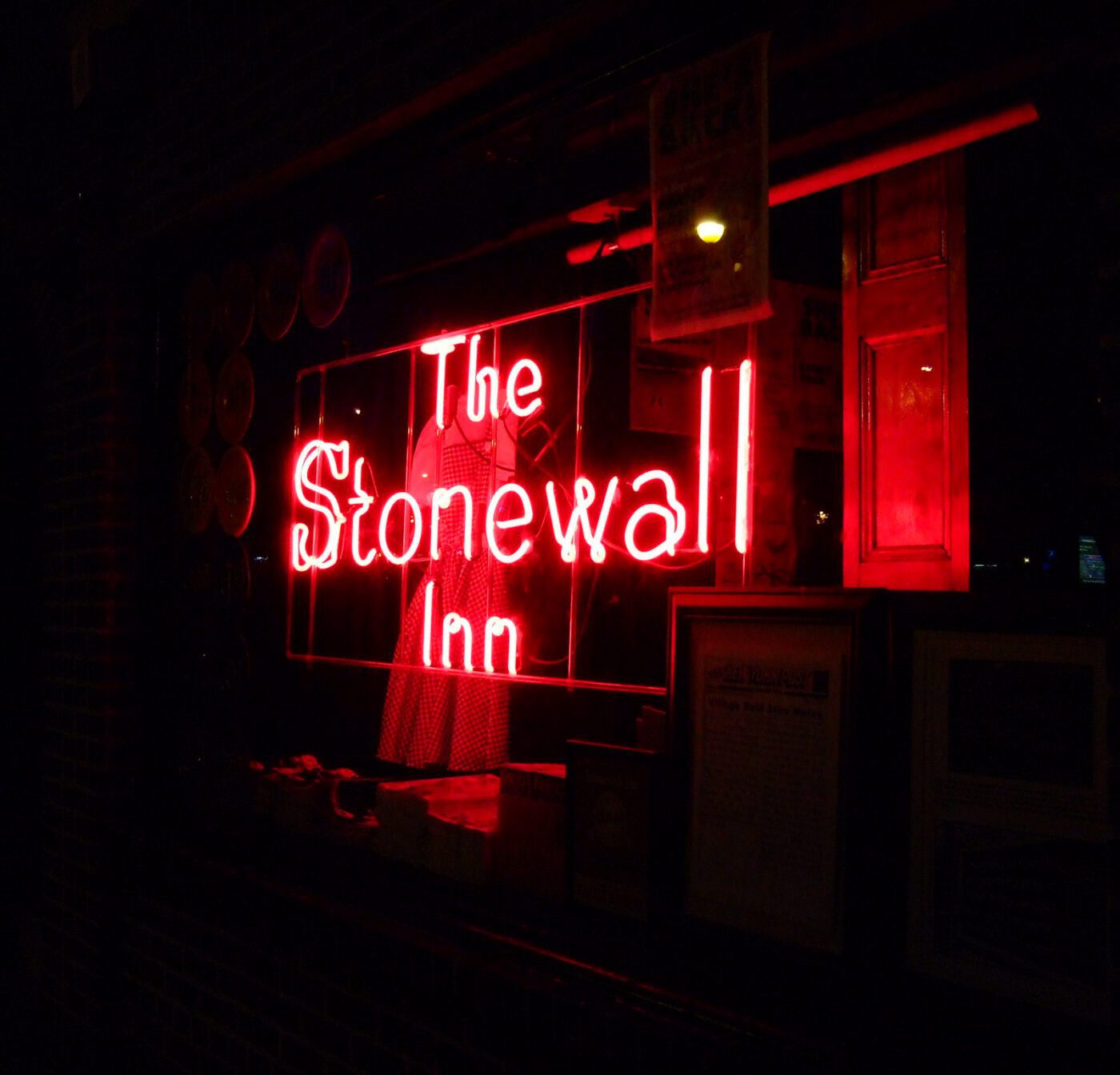 A neon sign glows red in a dark window, reading “The Stonewall Inn.” Some framed pictures and a dress are faintly visible inside.