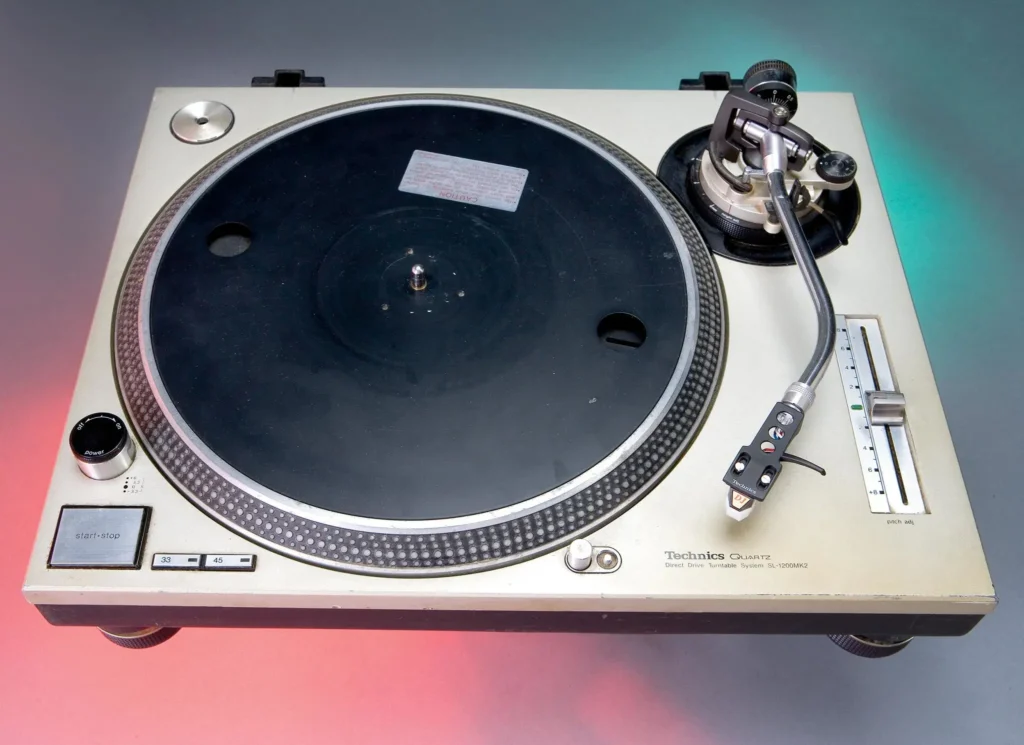 A vintage Technics Quartz turntable with a black platter, silver tonearm, and control buttons, set on a gray surface with red and green light accents.