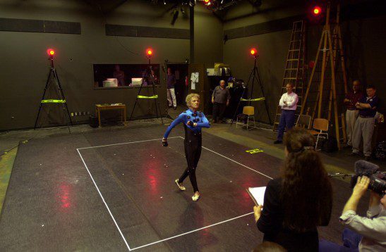 A person in a motion capture suit walks inside a marked area in a studio, surrounded by cameras and observers; red lights indicate recording equipment.