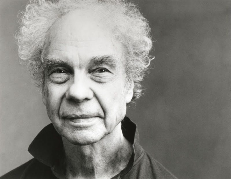Black and white portrait of an older person with curly white hair, slight smile, and wearing a dark collared shirt, looking directly at the camera against a plain background.