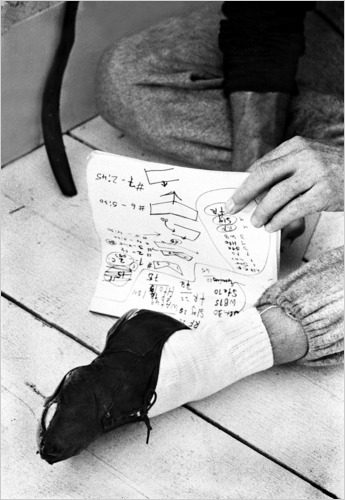 A person sitting on the floor points to handwritten notes with diagrams, while their sock-covered foot, partially wrapped with tape or fabric, rests on the wood planks nearby.