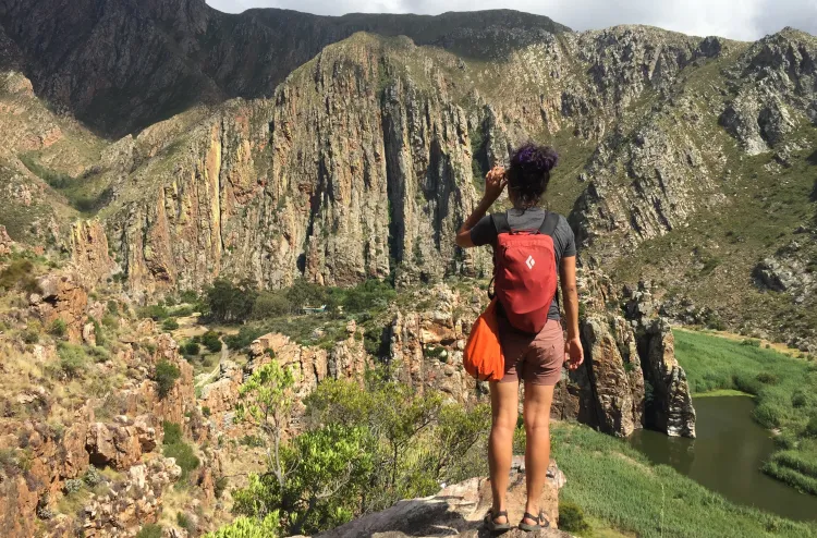 A person with a red backpack stands on a rocky ledge, overlooking a lush valley with steep cliffs, green vegetation, and a river below, surrounded by tall, rugged mountains.