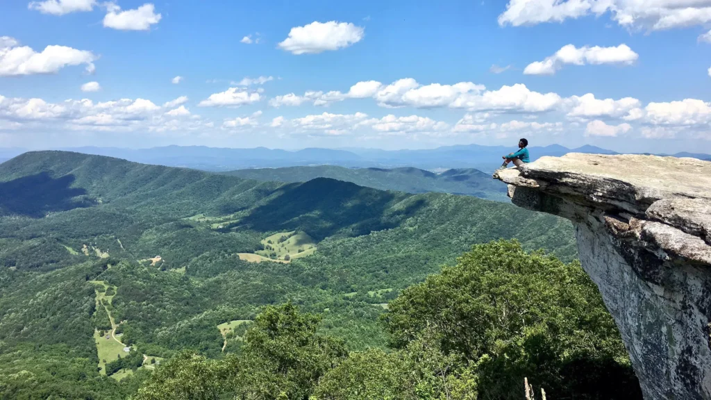 A person sits on a rocky cliff edge overlooking a vast, green mountainous landscape under a blue sky with scattered clouds.