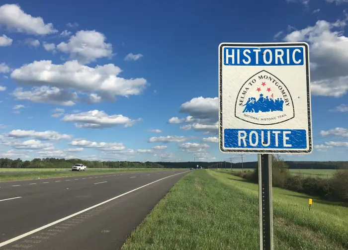 A blue and white road sign reads “Historic Route: Selma to Montgomery National Historic Trail” beside a highway under a blue sky with scattered clouds. A car is visible in the distance.