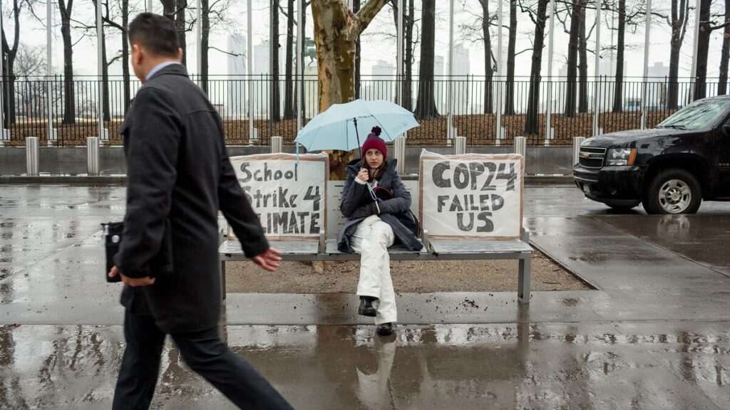 A person sits on a bench holding a blue umbrella between two protest signs reading “School strike 4 climate” and “COP24 failed us” as a person walks by on a rainy day.