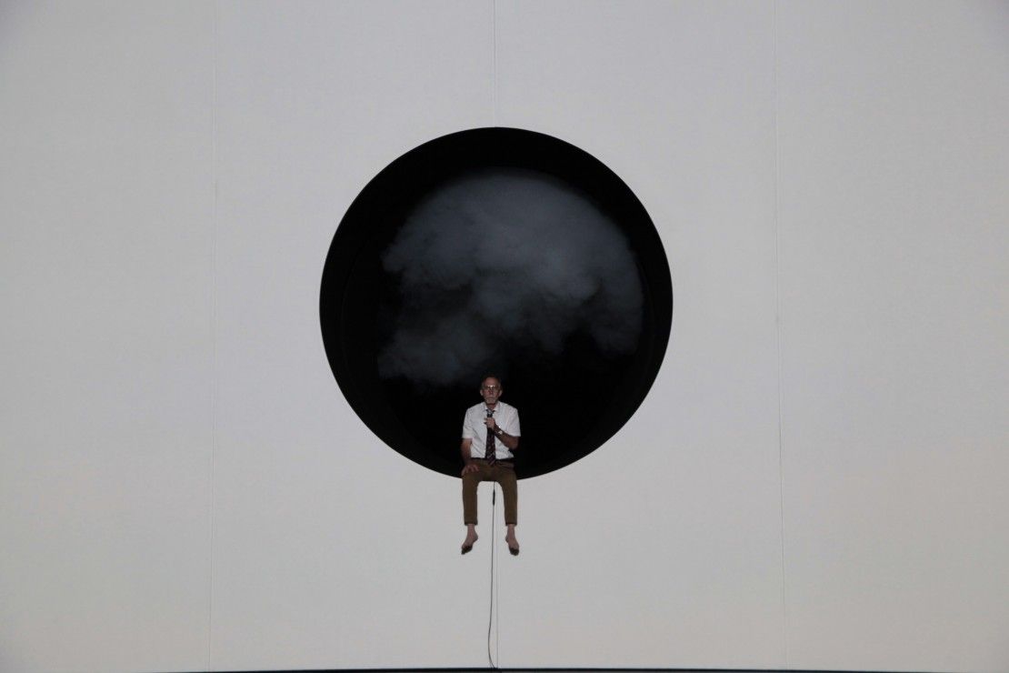 A person sits on the edge of a large black circular opening in a white wall, with smoke or mist inside the circle behind them.