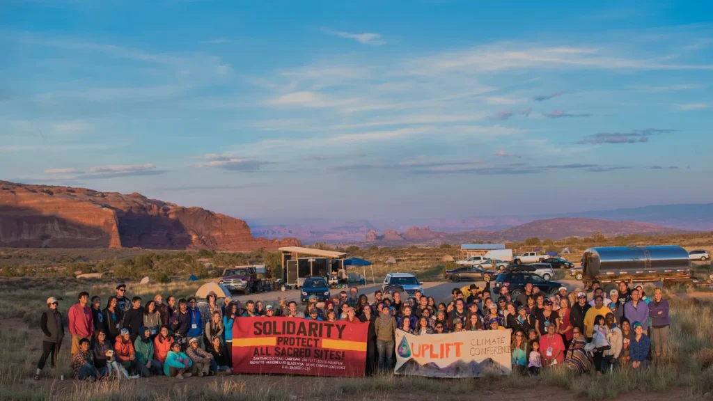 A large group stands in a desert landscape at sunset, holding banners reading “Solidarity Protect All Sacred Sites!” and “Uplift Climate Conference.” Cars and mesas are visible in the background.