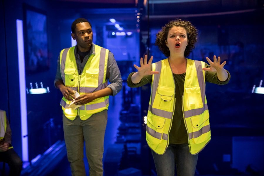 Two people wearing yellow safety vests stand indoors; one has hands raised, appearing to speak urgently, while the other looks on attentively. The background is dimly lit and slightly blurred.